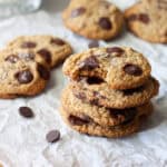 A stack of oat flour chocolate chip cookies on parchment paper with more in the background and a glass of milk to the side.