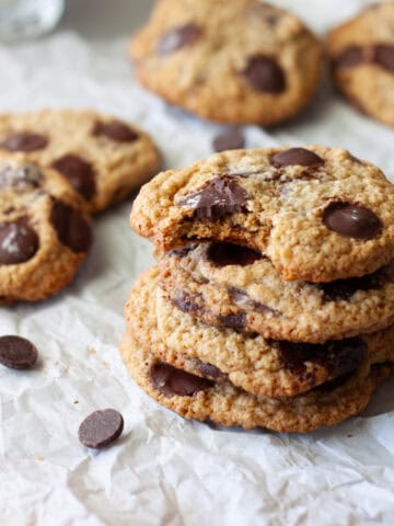 A stack of oat flour chocolate chip cookies on parchment paper with more in the background and a glass of milk to the side.