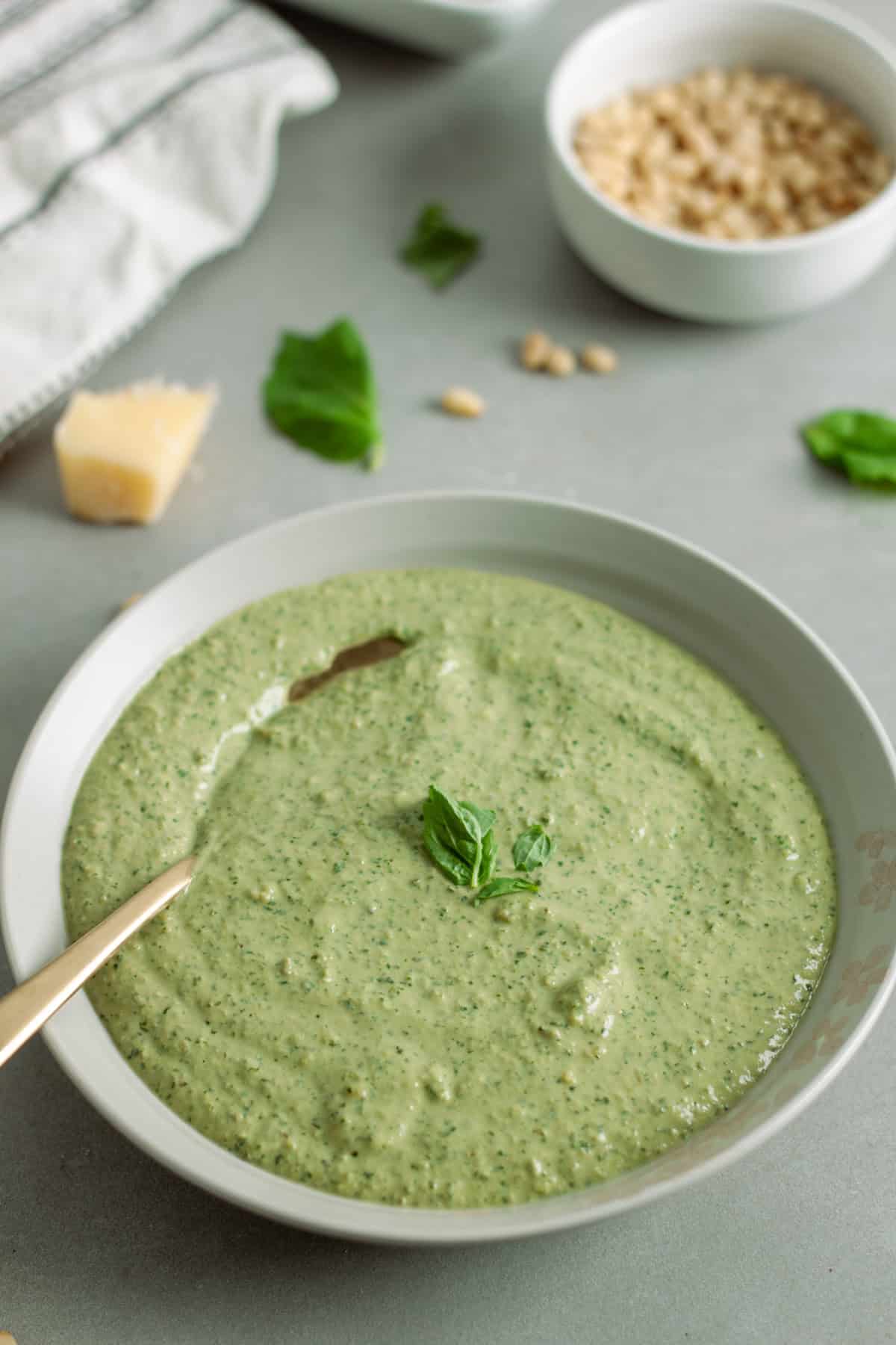 Ricotta pesto sauce in a bowl topped with fresh basil on a gray table with pine nuts in the background.