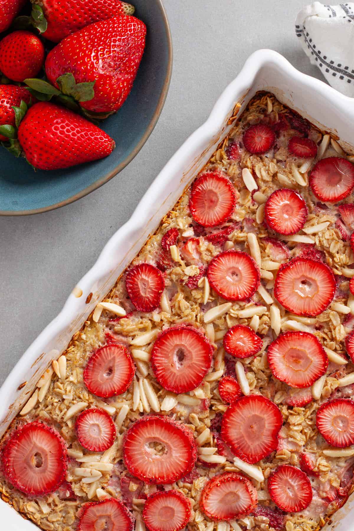 A close up of strawberry baked oatmeal in a white baking dish with a bowl of berries to the side.