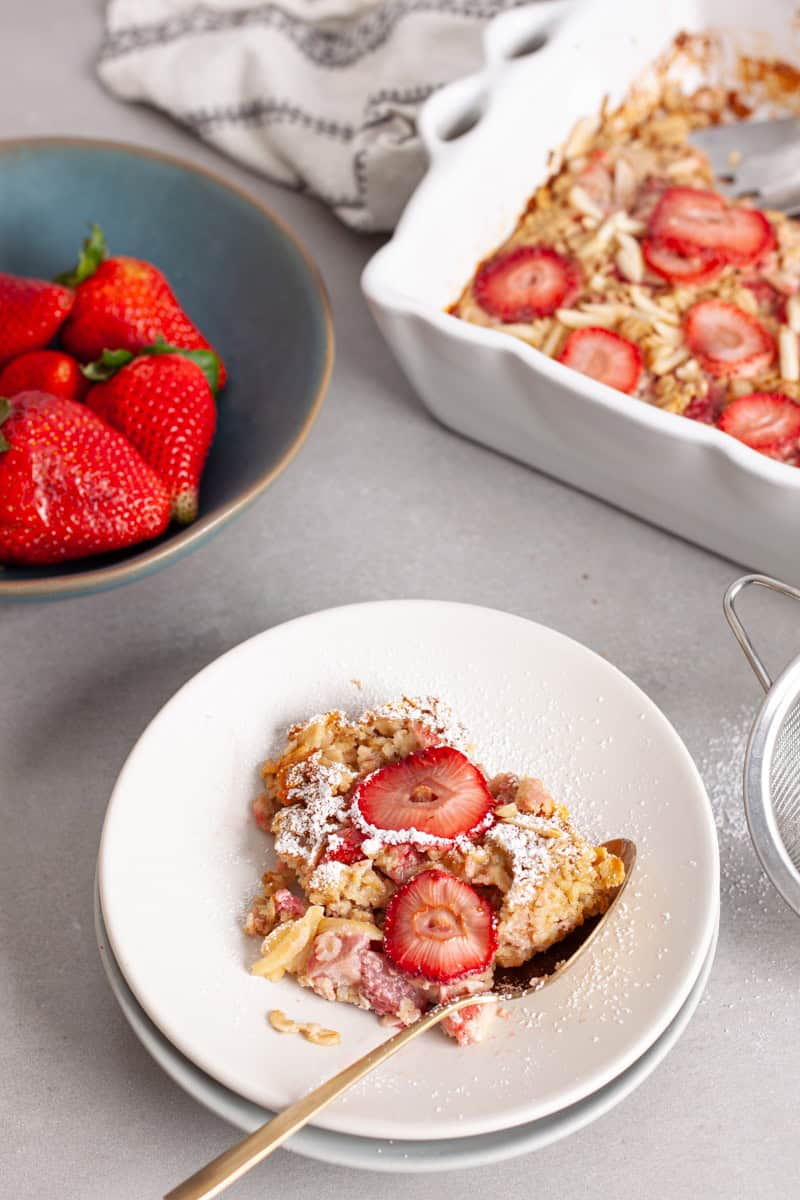 A serving of strawberry oatmeal on a small plate with a bowl of berries to the side on the baking dish in the background.