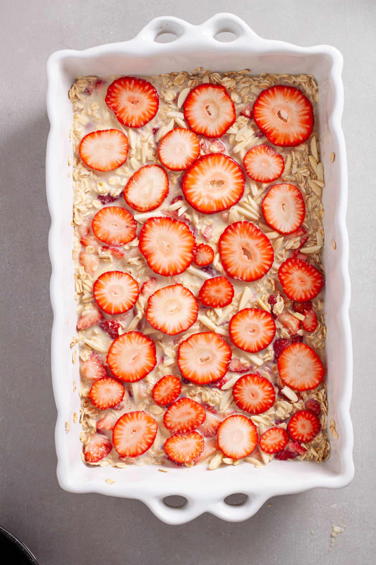 Uncooked baked oatmeal in a white baking dish topped with sliced strawberries and slivered almonds.