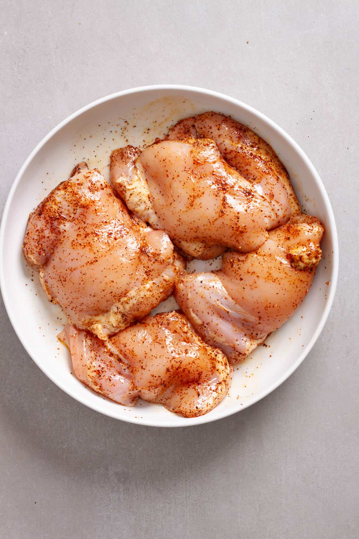 Chicken thighs tossed in a mixture of spices in a medium bowl.