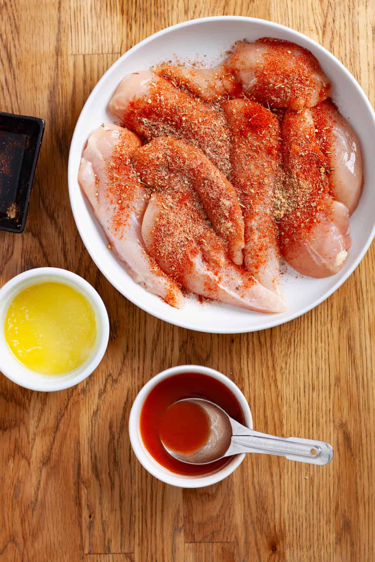 Chicken tenders seasoned with spices in a medium bow with melted butter and hot sauce to the side.