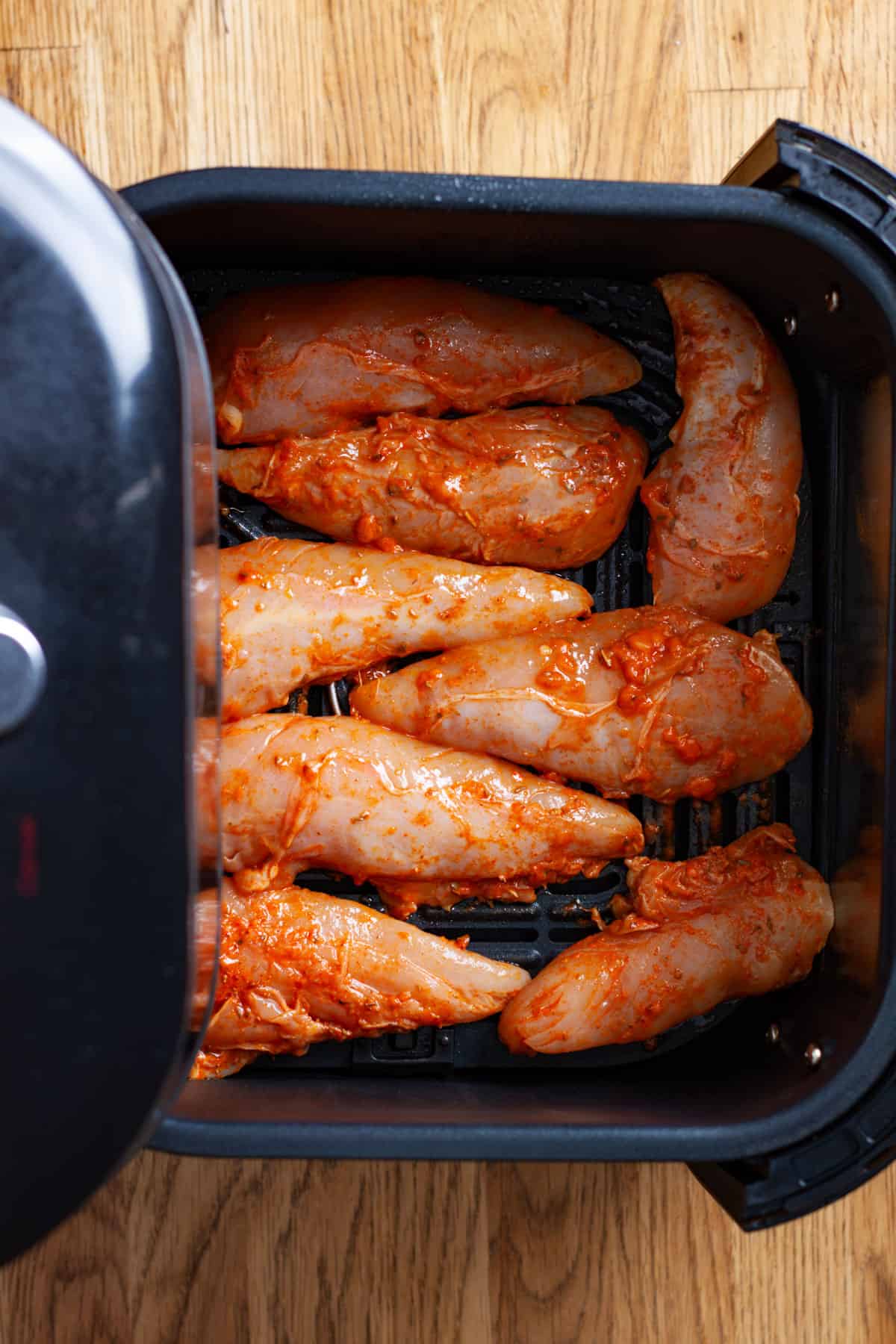 An air fryer basket with raw chicken tenders marinated in buffalo sauce ready to cook.