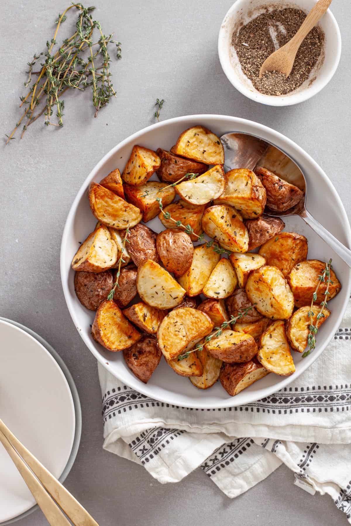 A serving bowl of crispy air fried red potatoes garnished with sprigs of fresh thyme and a bowl of black pepper to the side.
