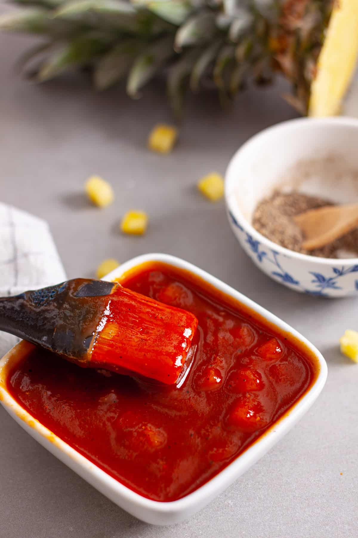 A small bowl of pineapple bbq sauce with a brush inside and a chopped pineapple in the background.