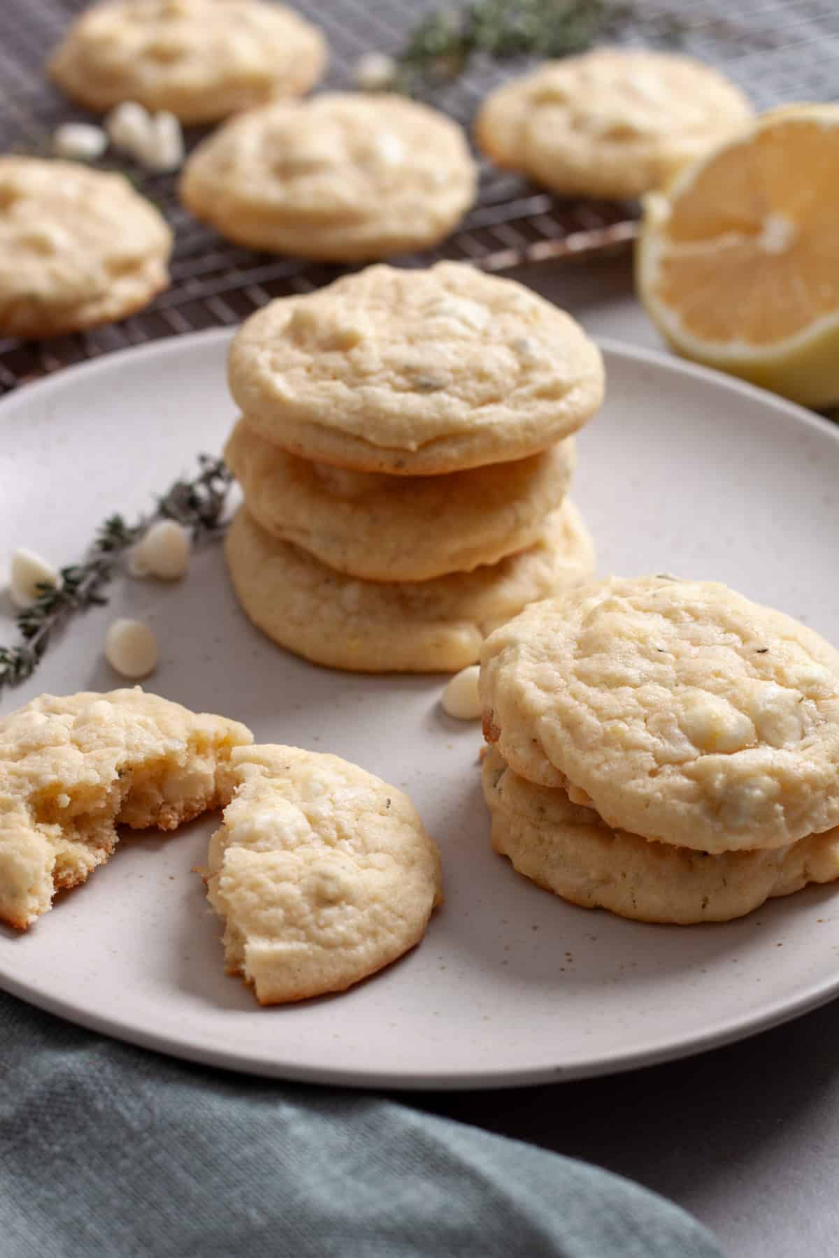 A plate with chewy lemon cookies with white chocolate and more in the background coolies on a rack.