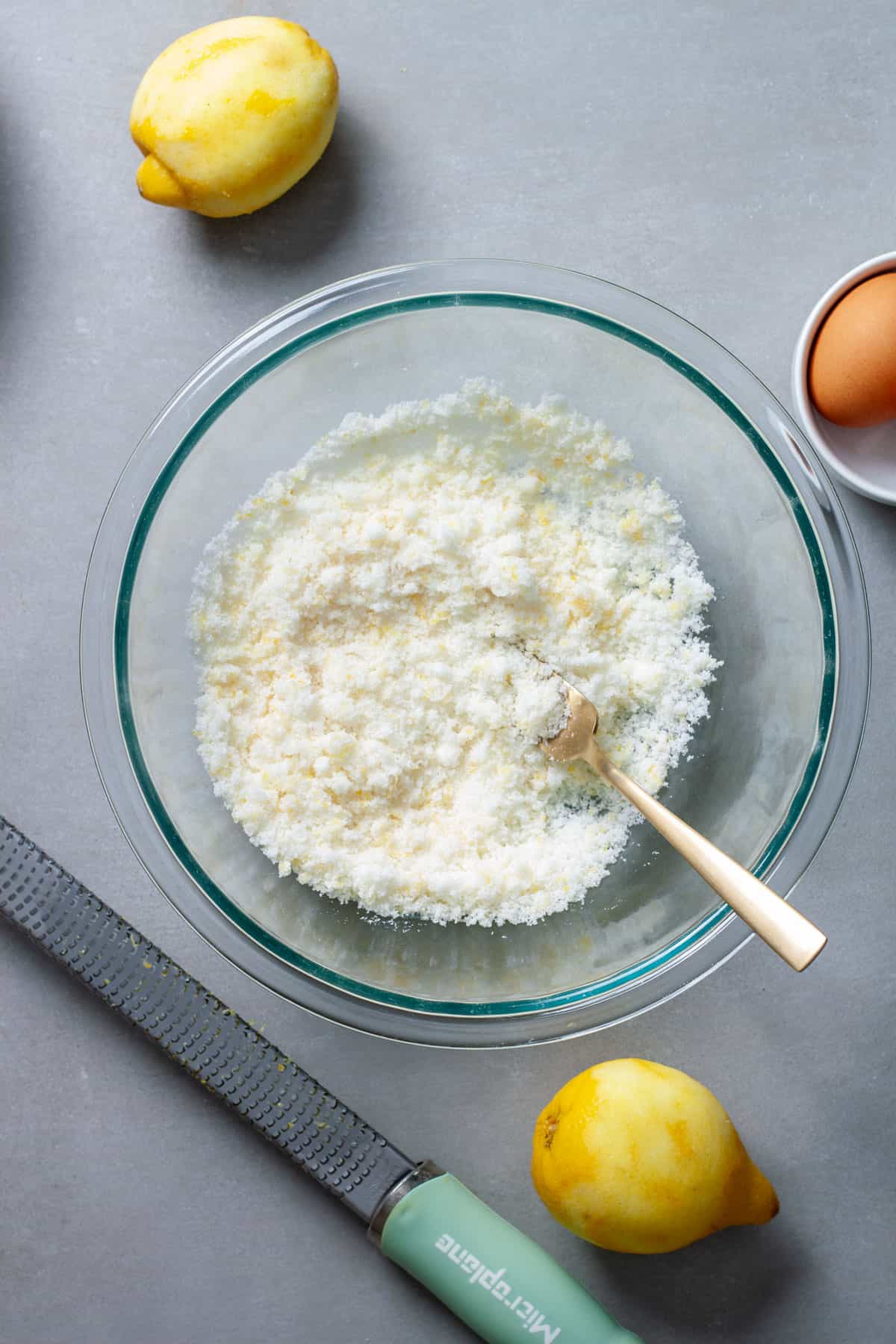 A fork mashing lemon zest into granulated sugar in a glass bowl on a gray table.
