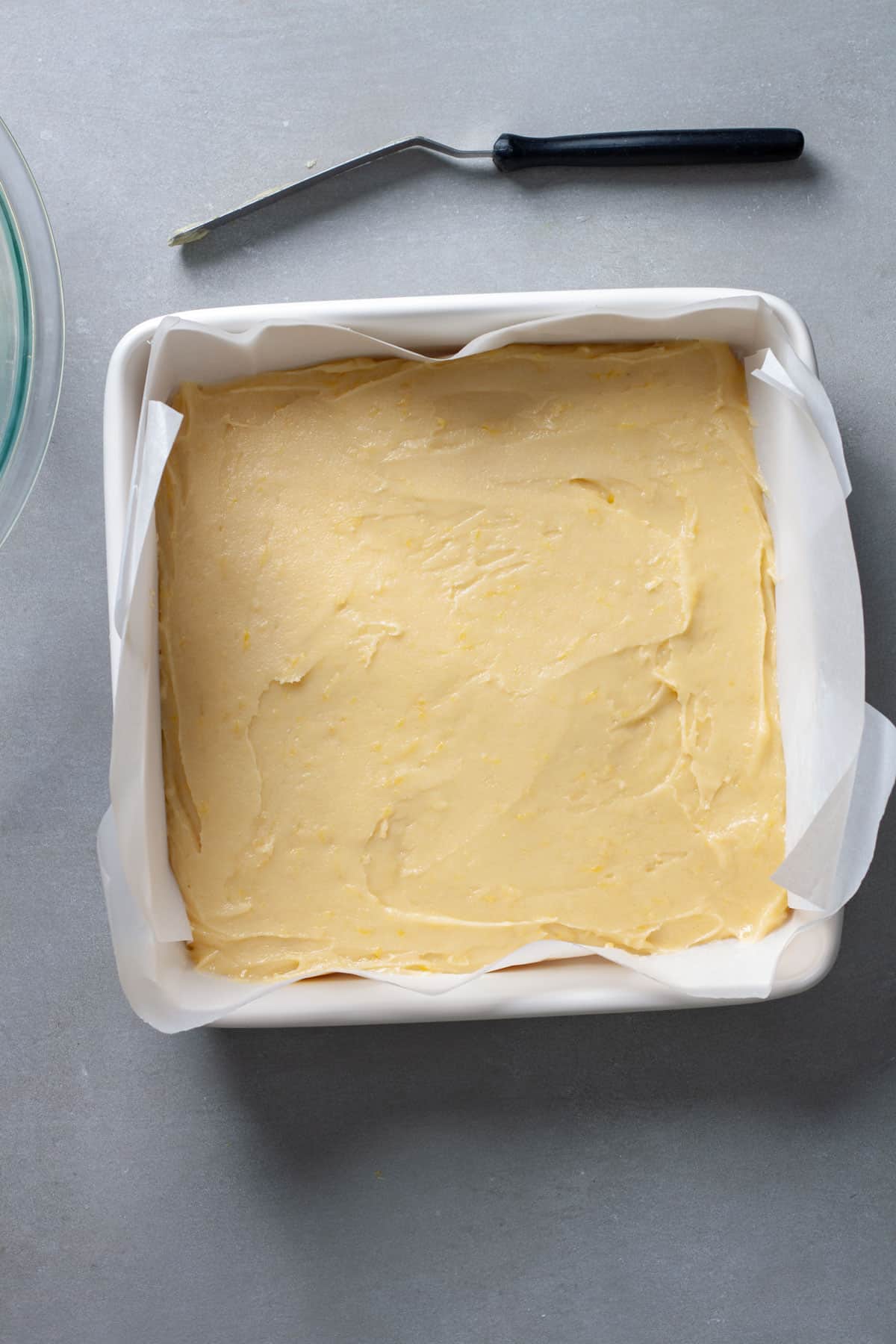 Lemon blondies unbaked in a parchment lined square baking dish on a gray table.