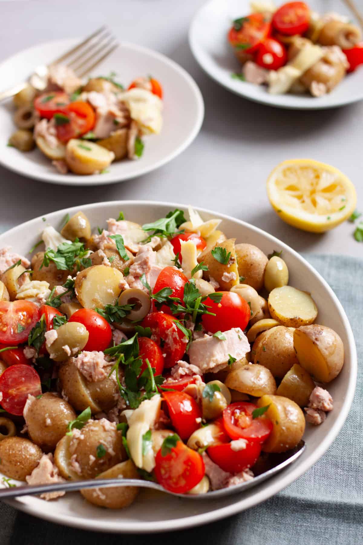A mixing bowl of potato salad with tuna and tomatoes and a serving in the background.