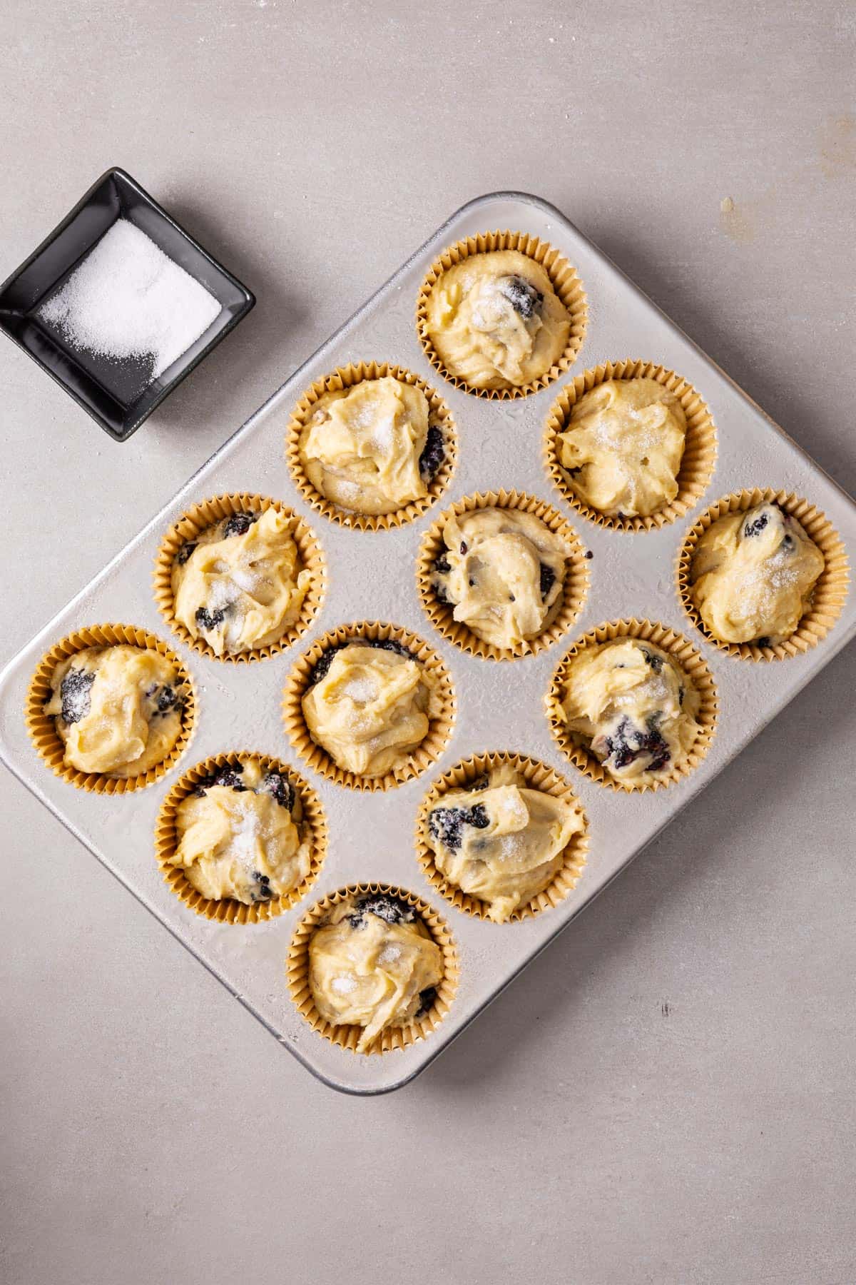 Blackberry muffin batter scooped into muffin cups with a small bowl of sugar to the side.
