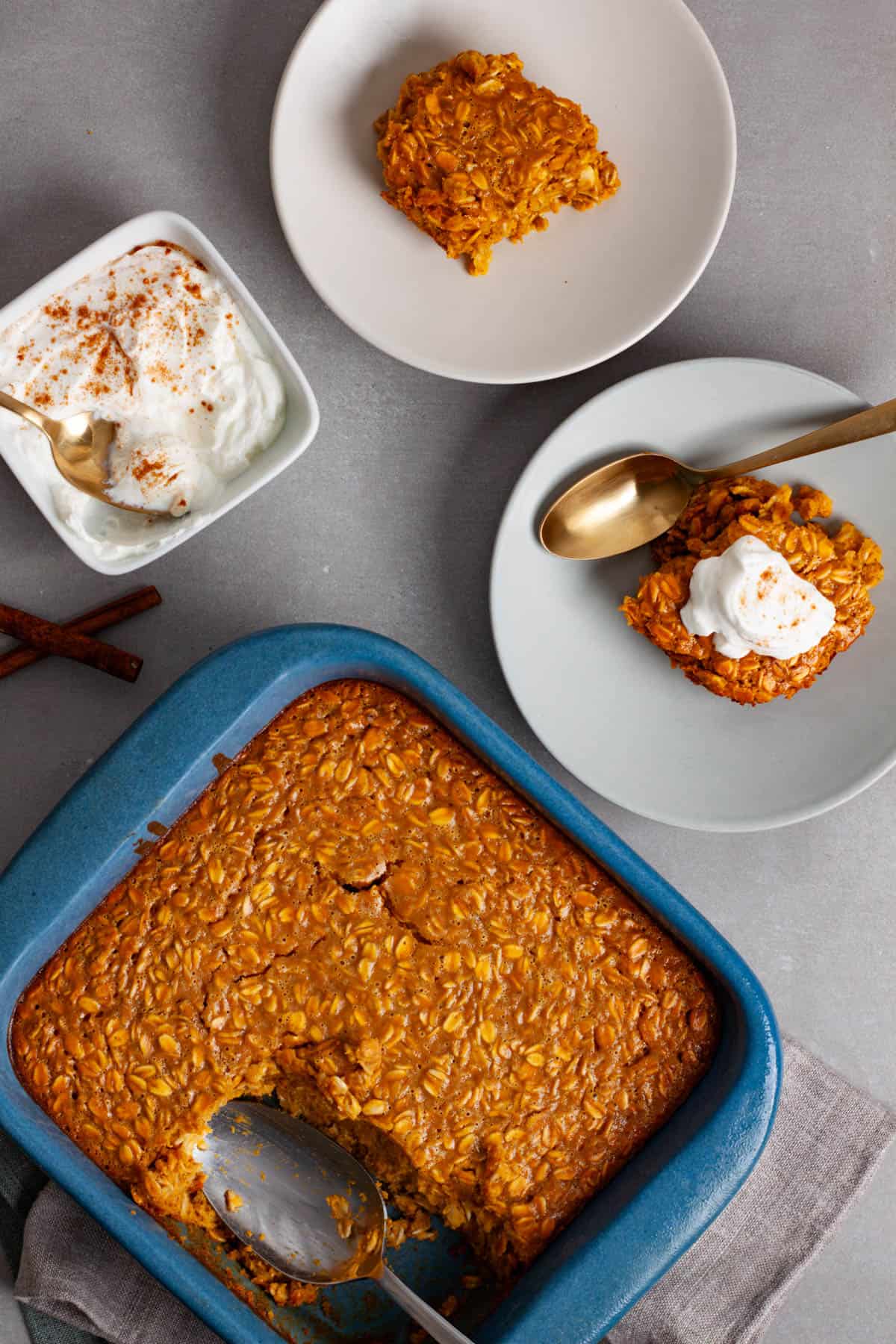 A gray table with pumpkin pie baked oatmeal n a baking dish and a couple servings to the side along with spiced yogurt to the other side.