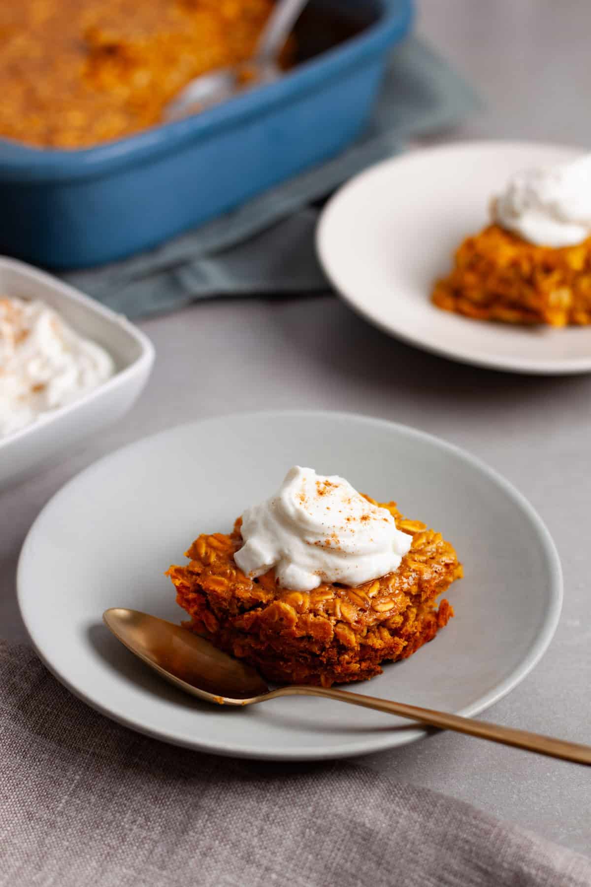 A portion of pumpkin baked oatmeal on a small plate with a dollop of spiced yogurt on top and more baked oatmeal in the background.