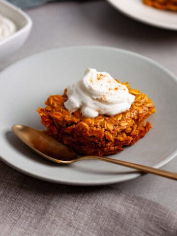 A portion of pumpkin baked oatmeal on a small plate with a dollop of spiced yogurt on top and more baked oatmeal in the background.