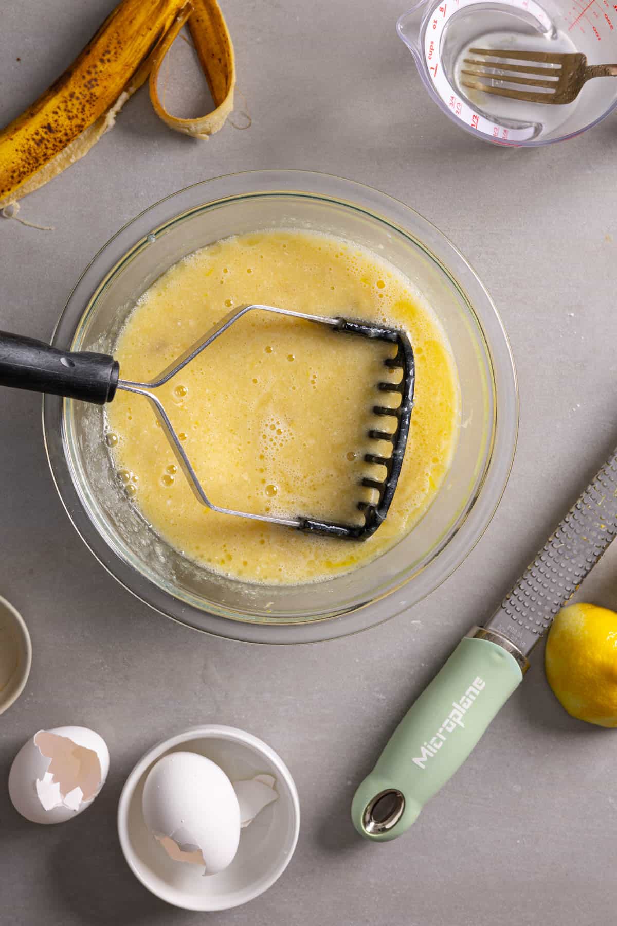A potato masher in a bowl with bananas and wet ingredients for muffins.