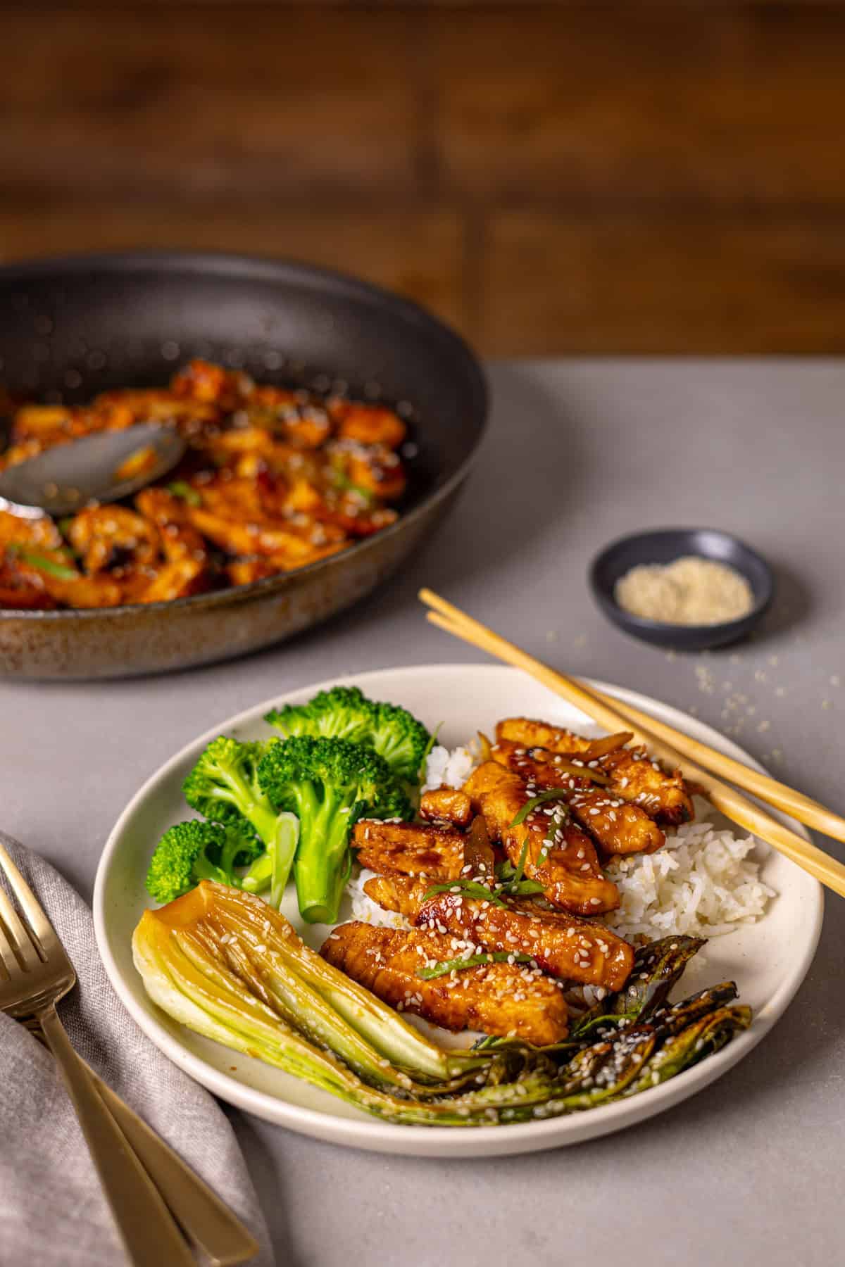 A serving of honey ginger chicken with rice and broccoli on a small plate and a skillet in the background with more chicken.