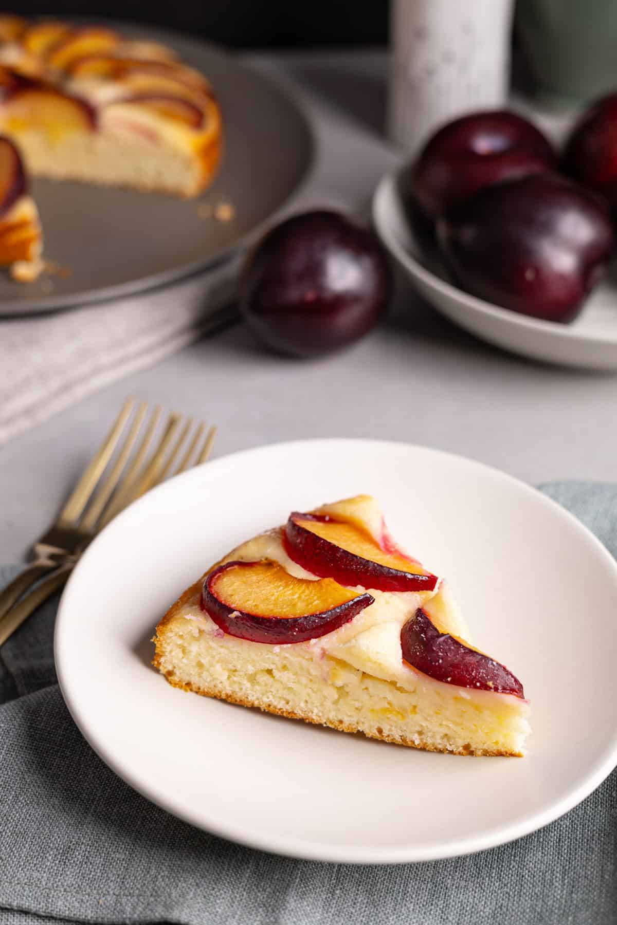 A slice of plum yogurt cake on a small plate with plums in the background.