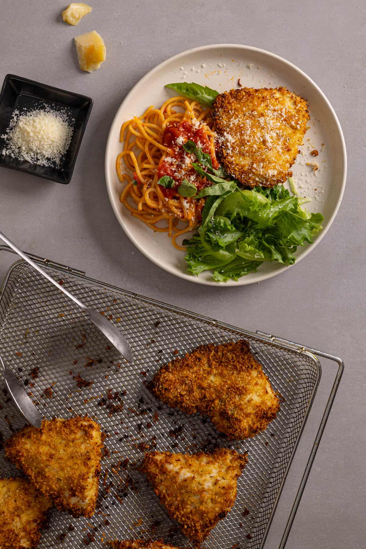 An air fryer basket with parmesan chicken and a plate with a serving of the chicken to the side.