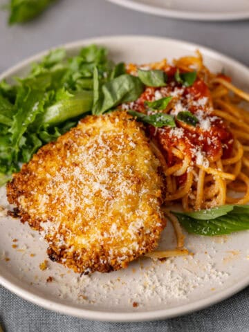 A plate with crispy air fryer parmesan crusted chicken, spaghetti, tomato sauce, and salad on a gray countertop.