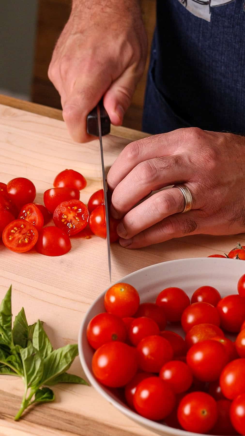 Cherry tomatoes getting cut in half on a wooden cutting board.