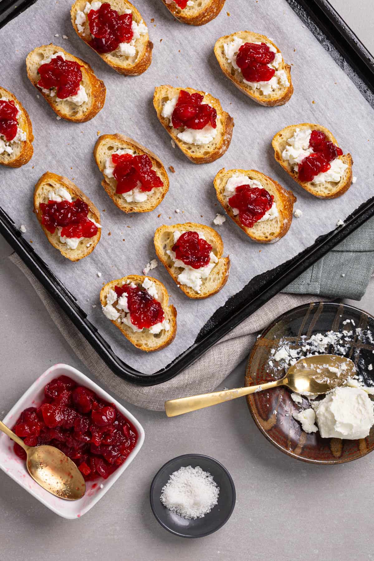 Candied cranberries spread onto slices of toasted baguettes with goat cheese on a parchment-lined baking sheet.