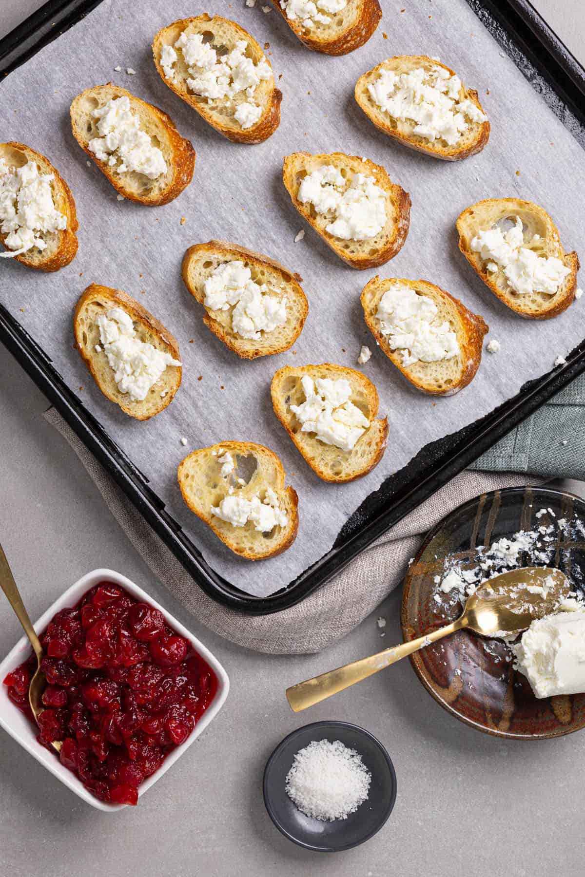 Goat cheese spread on a slices of toasted baguette on a parchment-lined baking sheet.