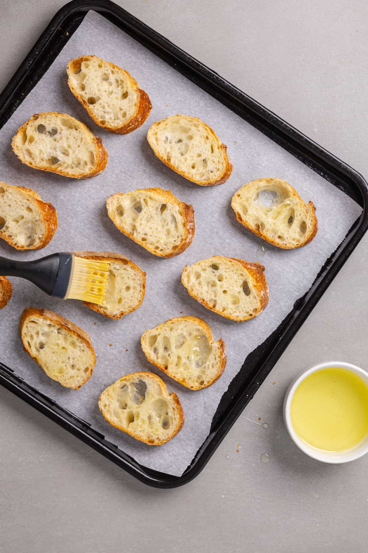 Slices of baguettes on a parchment lined baking sheet getting brushed with olive oil.