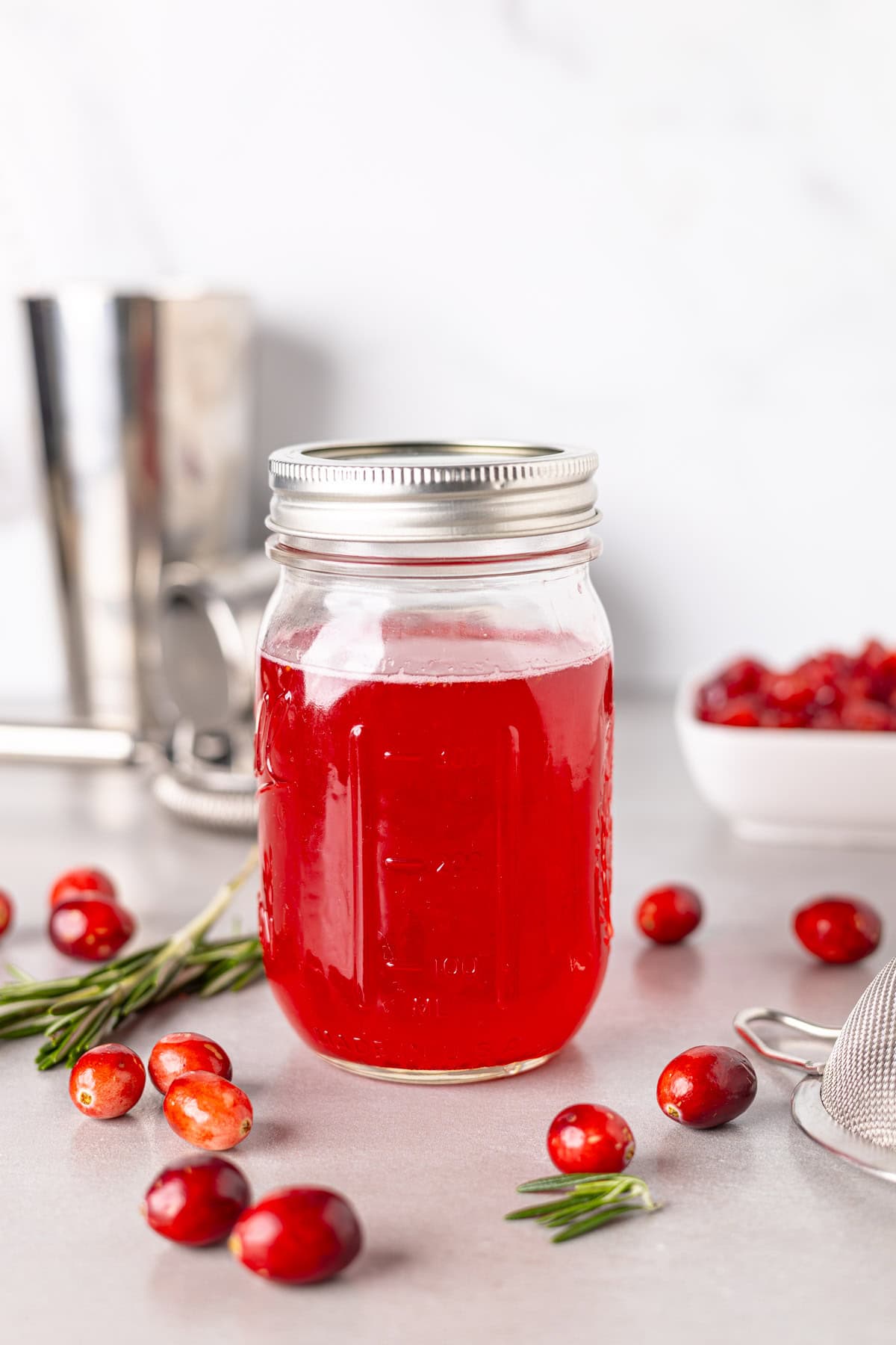 A mason jar with bright red cranberry simple syrup and fresh cranberries spread around the table.