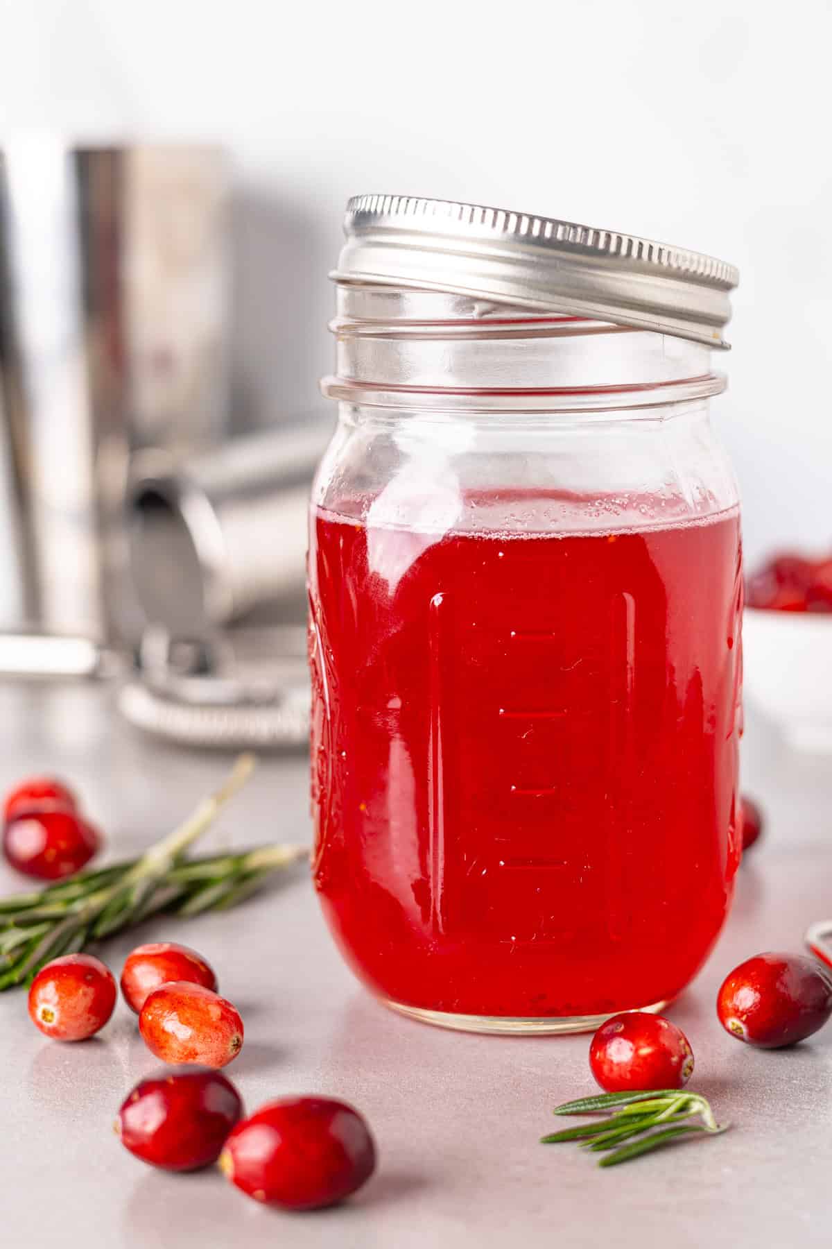 A close up shot of a jar with cranberry simple syrup on a gray table with fresh cranberries on the table.