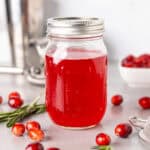 A mason jar with bright red cranberry simple syrup and fresh cranberries spread around the table.