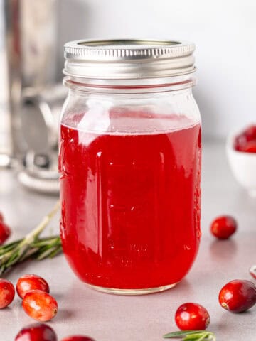 A mason jar with bright red cranberry simple syrup and fresh cranberries spread around the table.