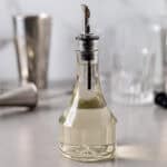 A glass bottle with a sprout on top filled with a clear simple syrup liquid and cocktail mixing tools in the background.