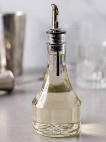 A glass bottle with a sprout on top filled with a clear simple syrup liquid and cocktail mixing tools in the background.