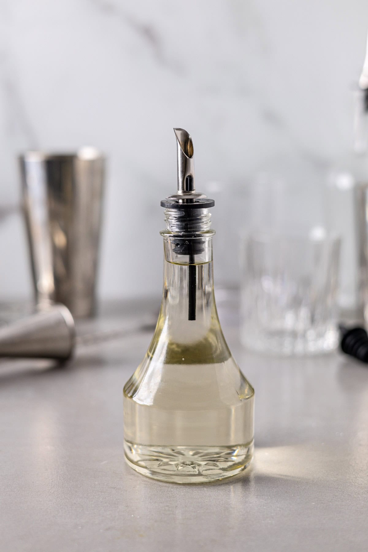 A glass bottle with a sprout on top filled with a clear simple syrup liquid and cocktail mixing tools in the background.