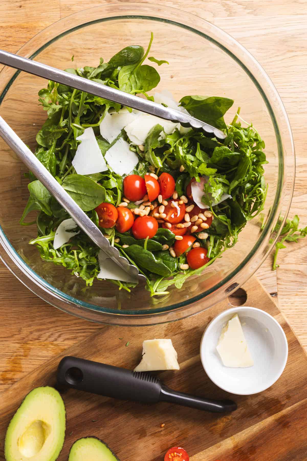 A spinach and arugula salad with tomatoes, pine nuts, and aged provolone getting tossed in a large mixing bowl.