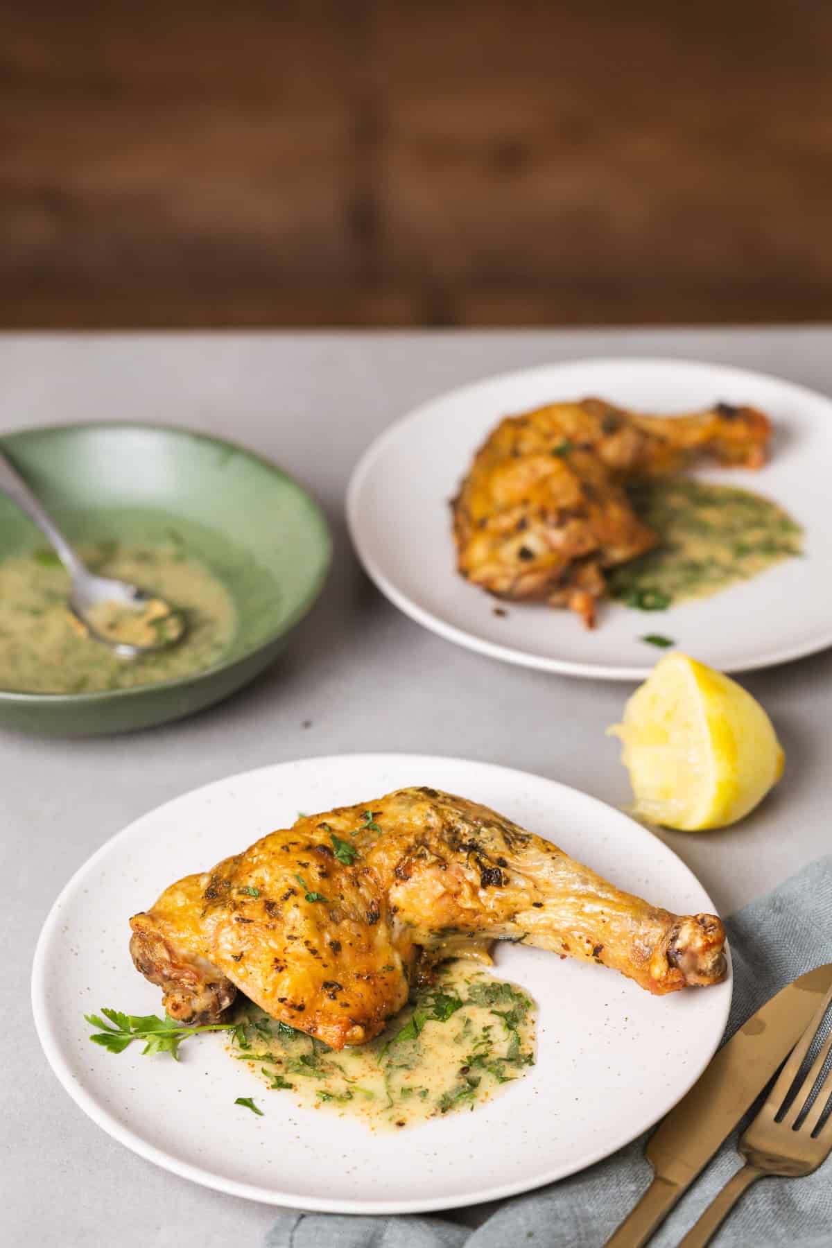 A serving of golden brown roasted chicken leg quarters with an herb sauce on a gray table with more chicken in the background.