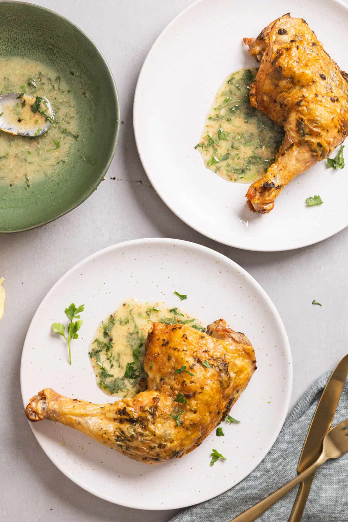 Roasted chicken leg quarters garnished with an herb and lemon sauce on two small white plates.