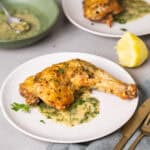 A serving of golden brown roasted chicken leg quarters with an herb sauce on a gray table with more chicken in the background.