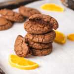 A stack of chewy chocolate and orange cookies on a parchment-lined baking sheet with slices of oranges as a garnish.