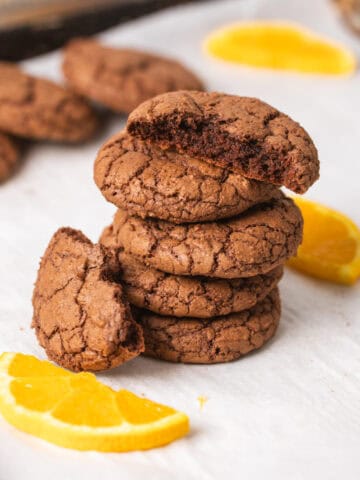 A stack of chewy chocolate and orange cookies on a parchment-lined baking sheet with slices of oranges as a garnish.