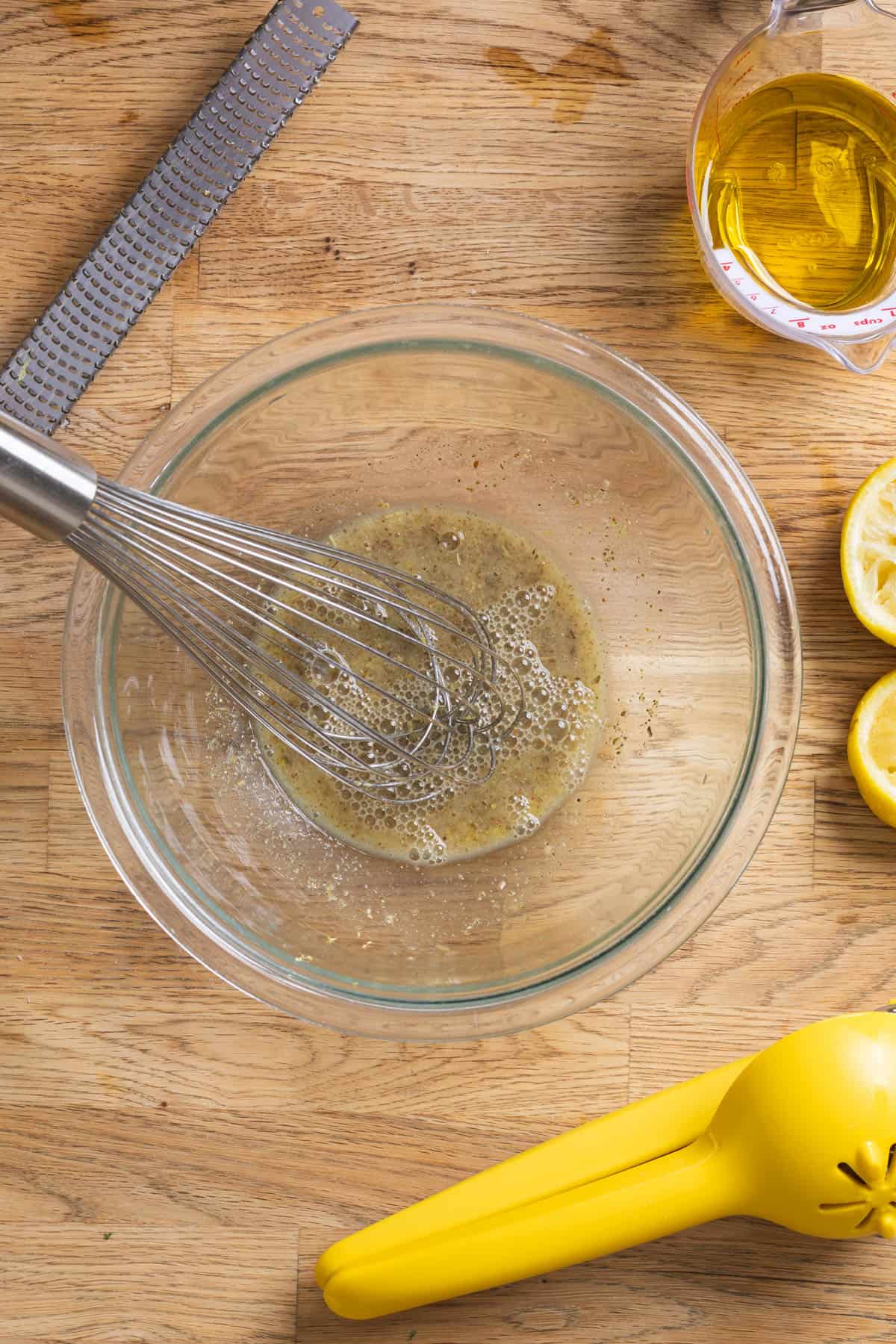 A whisk blending together ingredients for a lemon Dijon dressing in a glass bowl on a butcherblock countertop.
