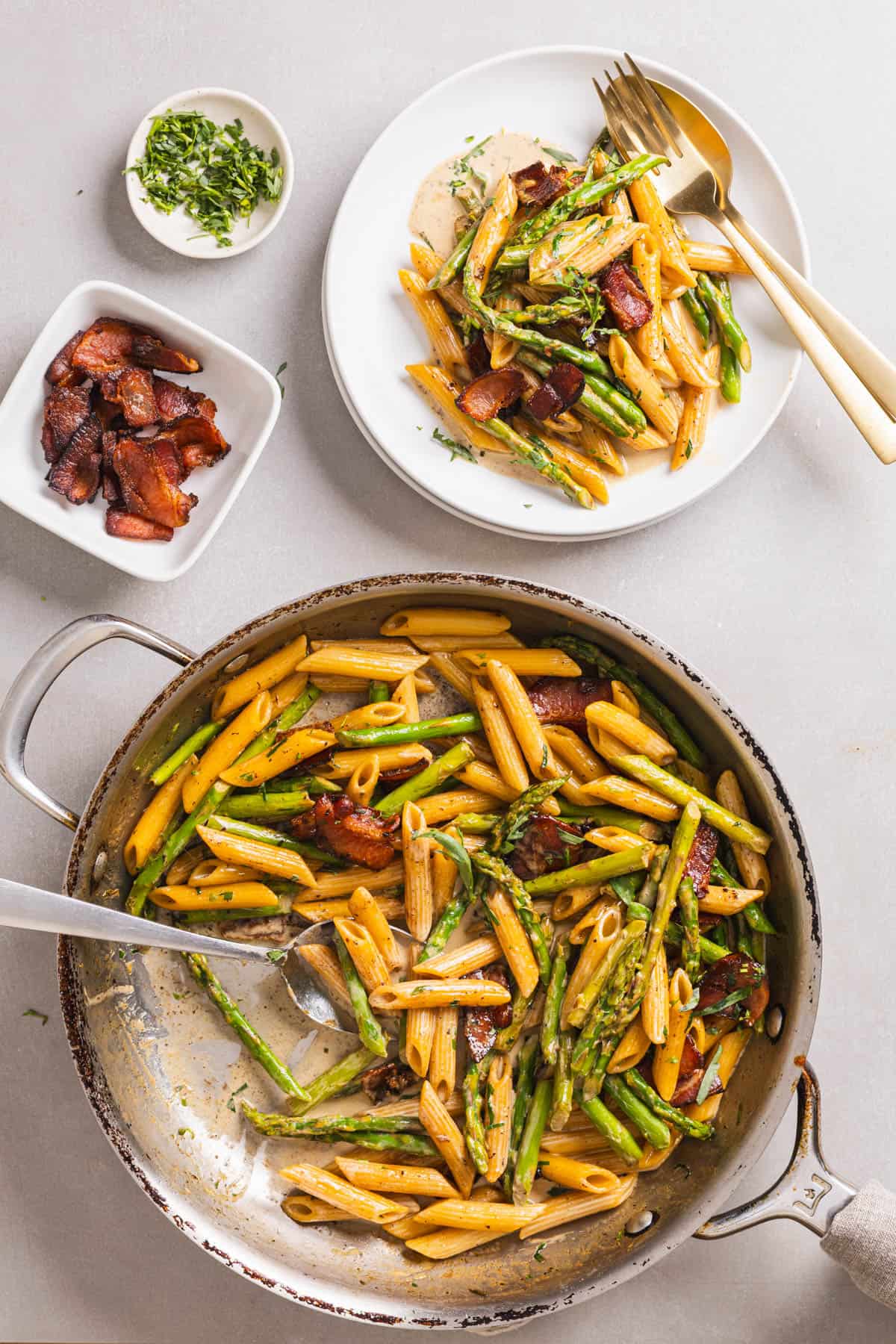 A skillet with bacon and asparagus pasta with a large spoon and a small white plate with the pasta to the side.