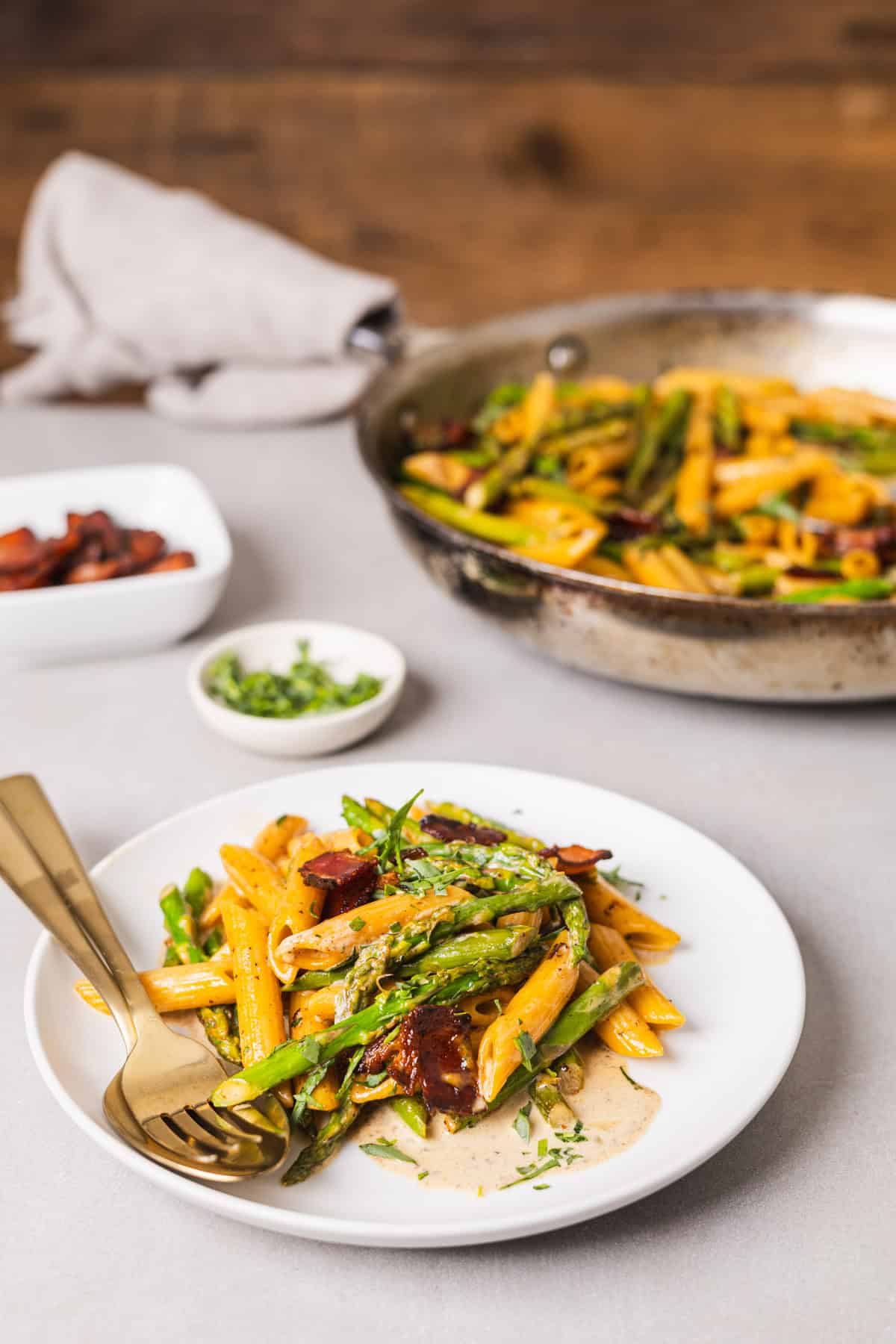 A white plate with bacon and asparagus pasta with a skillet with more pasta in the background.