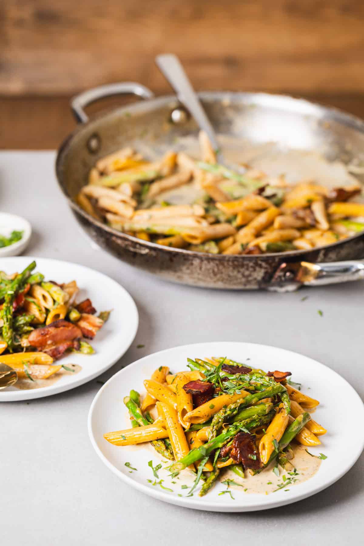 Two small plates with bacon and asparagus pasta on a gray table and a skillet with more pasta in the background.
