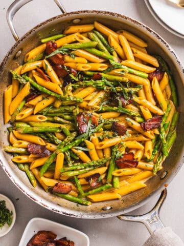 A close-up shot of a skillet with bacon and asparagus pasta garnished with fresh tarragon.