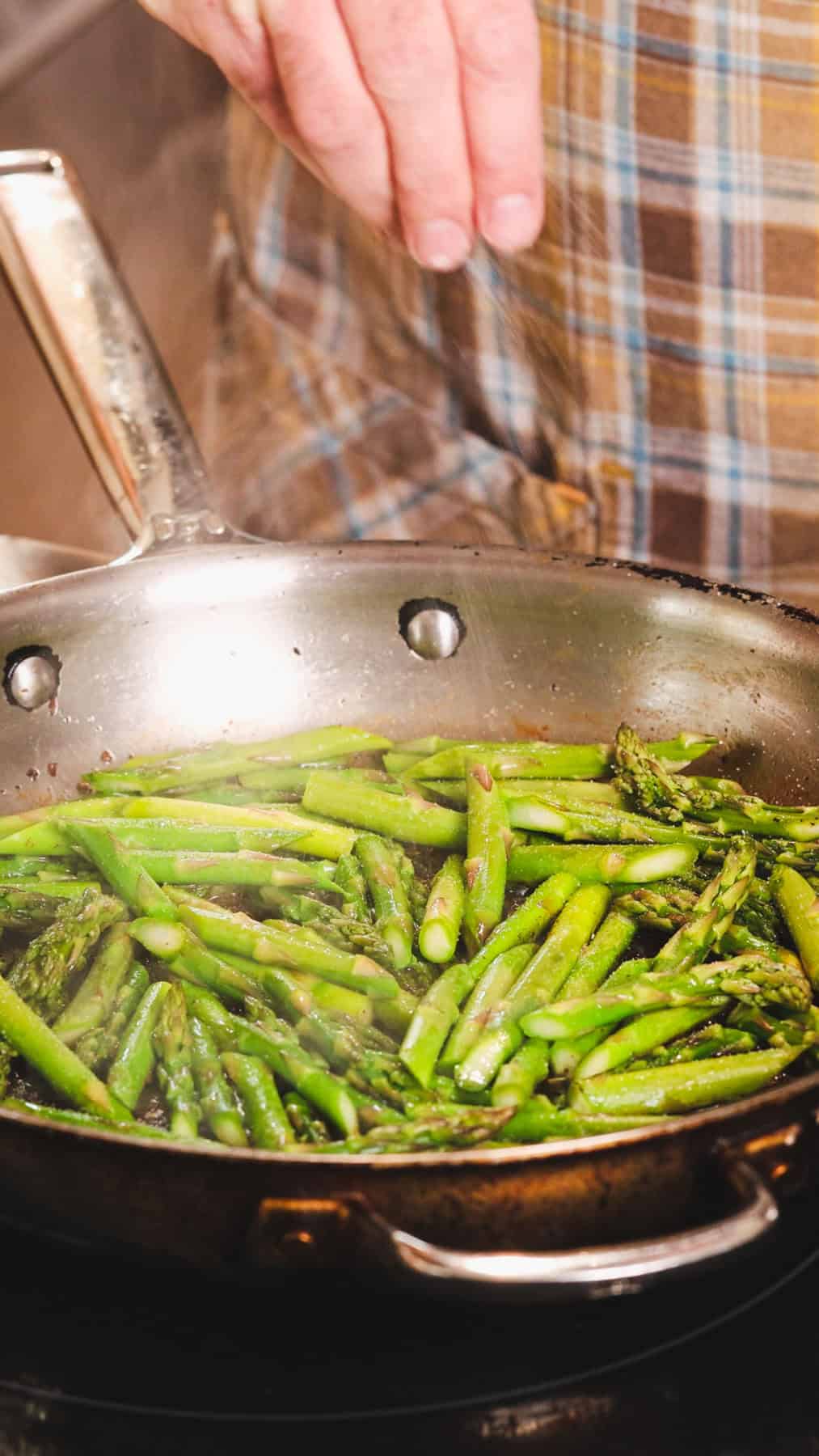 Asparagus cut into 2-inch pieces cooking in a large skillet and a hand seasoning it with salt.