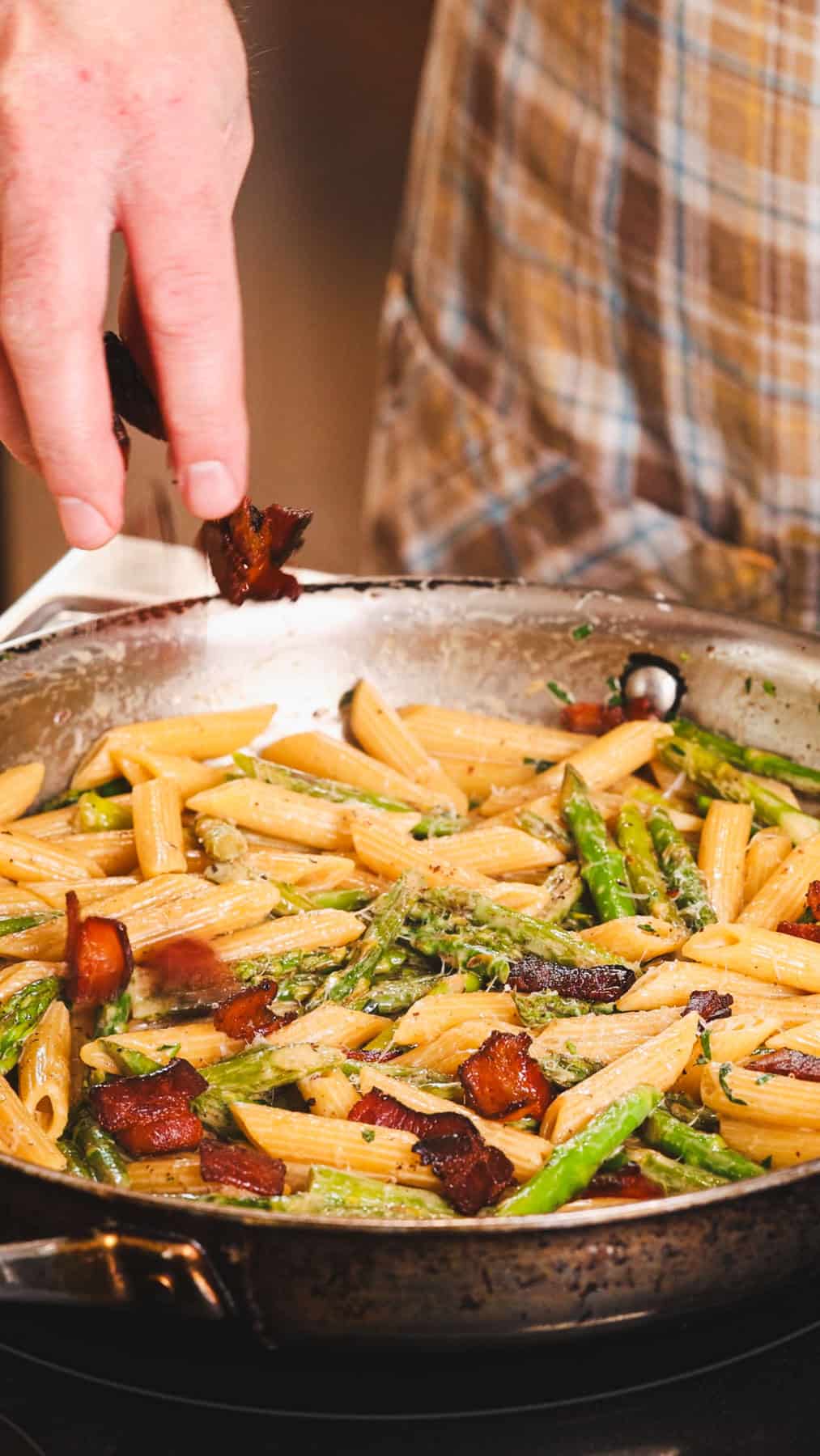 Crispy cooked bacon getting sprinkled into a skillet with pasta and asparagus and a light cream sauce.