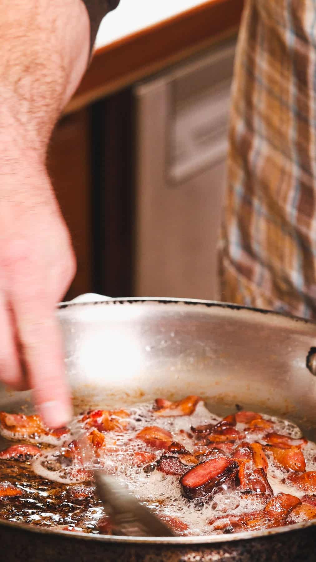 Chopped bacon cooking in a large skillet and getting stirred by a large metal spoon.
