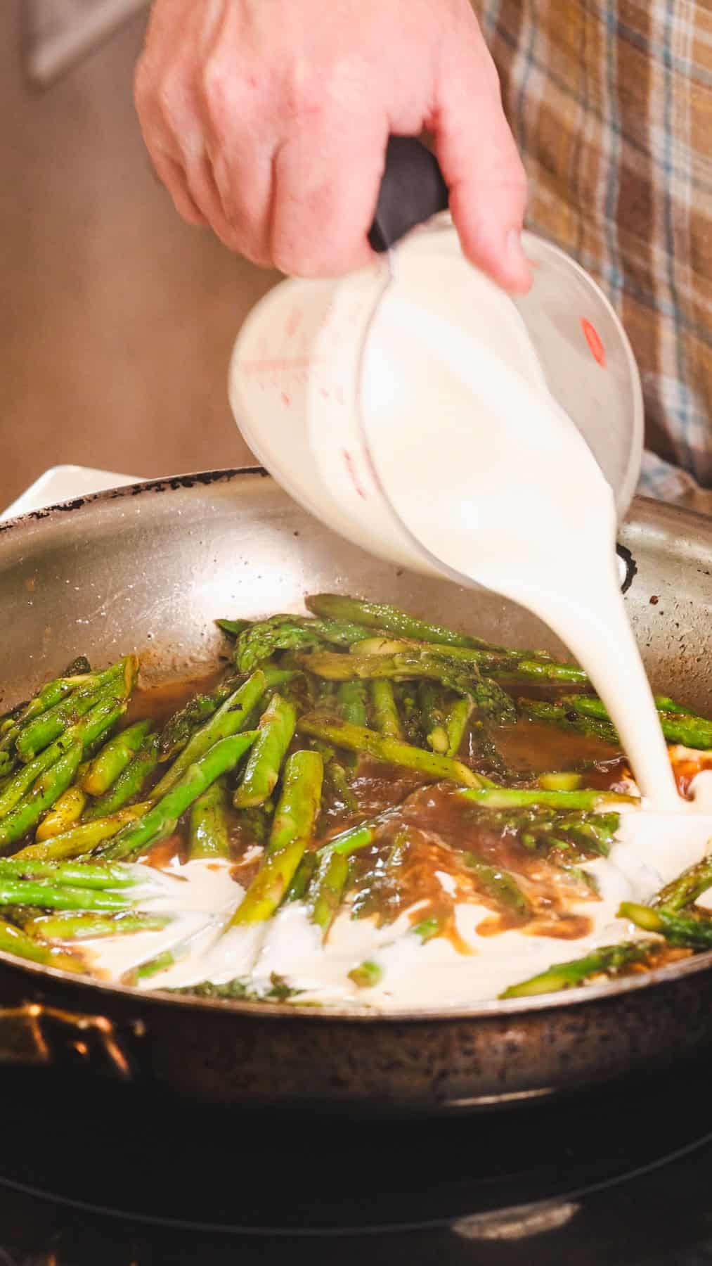 Heavy cream getting poured into a skillet with asparagus for a bacon and asparagus pasta.